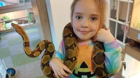 Gareth Costellow's daughter with a snake around her neck at the reptile centre and looking happy. She is wearing a colourful jumper and the snake is light and dark brown. There are animal enclosures in the background.