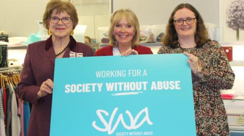 Three women stand in a charity shop holding a large blue sign saying 'Working for a society without abuse'
