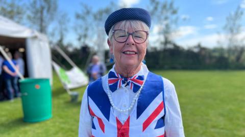A woman wearing a Union flag bow tie and waist coat. She has a pearl necklace and a hat. She also has glasses and short grey hair. 