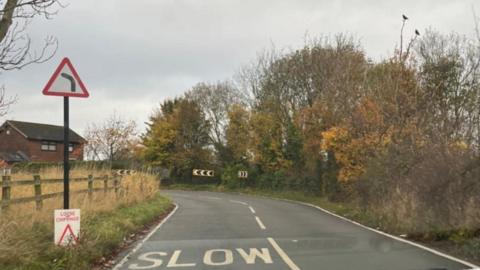 A harsh bend on a country road, surrounded by trees and vegetation. The road markings read slow, and there are black and white arrows showing the severe bend in the road. By a fence, there is a sign that reads 'loose chippings'