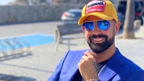A man tanned smiling at the camera wearing aviator sunglasses a yellow Dsquared2 baseball cap and electric blue jacket and black T-shirt. He has a trimmed black beard. He is sat near a swimming pool which is out of focus behind him.