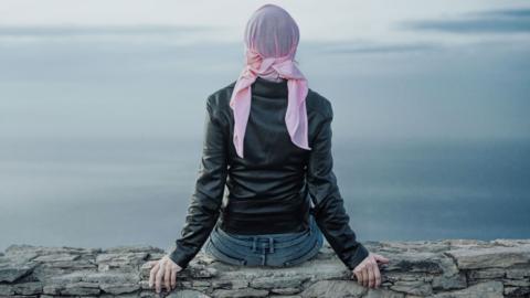 A shot of a woman from behind. She is wearing a pink headscarf and black leather jacket andsitting on a wall looking out to sea
