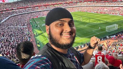 Bhavs, a South Asian man at Arsenal's Emirates stadium. He is wearing a black vest top with a light and dark blue striped top underneath. His left hand is raised in a fist pump action. Behind him is a green football pitch with thousands of fans in the stadium around him.