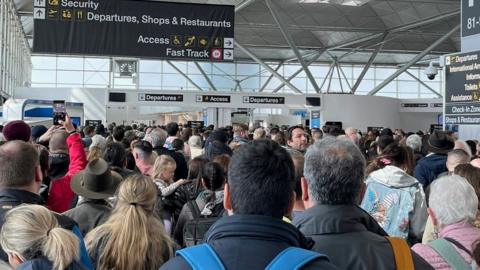 Crowd of people in terminal building at London Stansted Airport. Signs above directing to "departures"