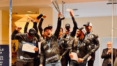 A group of men throw their hands into the air in celebration. The one in the middle is holding up silver trophy. They are all wearing baseball caps.