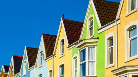 A row of closely spaced terraced houses viewed from a low angle, emphasizing their sloping roofs and upper floors, on a bright sunny day with a blue sky. The houses are painted in bright, contrasting pastel colours - including light blue, pale yellow and a vivid lime green.