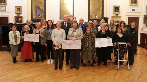 A group of men and women standing inside a parish hall in St Helier, Jersey, during a cheque presentation ceremony. Three large cheques are being held by women in the group.