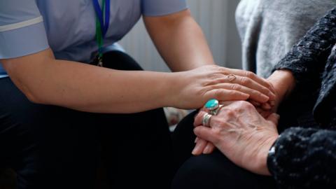 A close-up shop showing a person in a nurse uniform sat in front of an elderly person. The nurse has their hands over the elderly persons hands. 