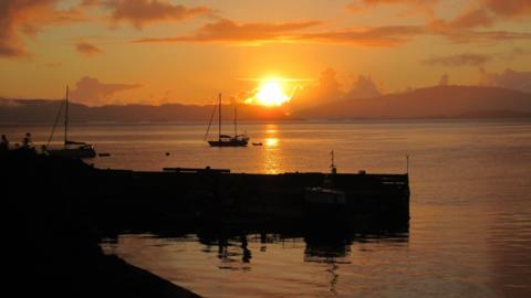 An orange sunset over a harbour, with sailing boats in the water.