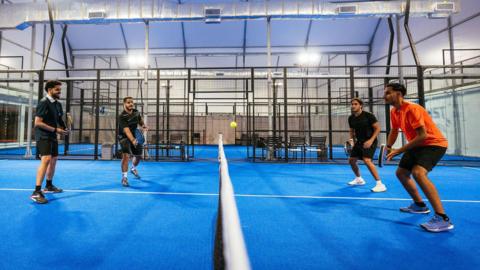 A padel court with a bright blue floor and cages surrounding the court. A doubles match is taking place, and the four men are playing, dressed in shorts and t-shirts. 