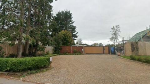 The image shows a piece of land opposite a farm. The sky is grey and there are trees growing on the left hand side. A house can be seen in construction on the right and a Union Jack can also be seen on top of the house.