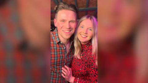 Man and woman both with clone hair smiling. The man is wearing a red checked shirt the woman is wearing a red top with black spots.