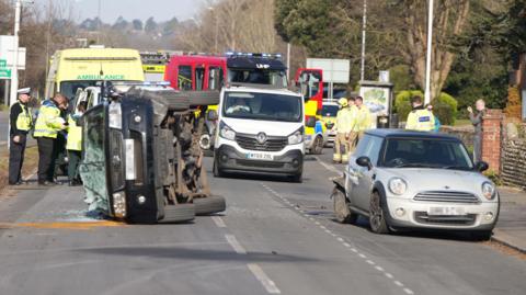 A black Suzuki vehicle on its right side in the road. Its windscreen is smashed. To its right is a silver Mini. There are various emergency workers and vehicles in the background