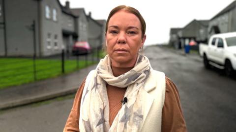 A woman is facing the camera. She has brown hair that is tied up. She's wearing a light brown long-sleeve top and a cream scarf with grey stars. The background is blurred but shows a street of dark grey houses. There is a white truck to the side. 