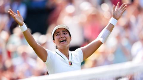 Filipina tennis player Alexandra Eala raises his arms after a Wimbledon match, net in front of her and a blurred image of the crowd in the background