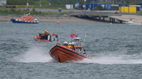 Three vessels on a body of water with a jetty behind them. There are two smaller boats in the foreground, with one RNLI orange craft heading out from shore, creating a spray. There are several people on board wearing white helmets and lifejackets. 