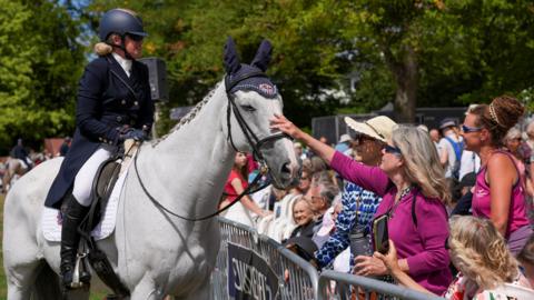 A woman on a white horse, wearing smart riding clothes, stands near a fence at the Taunton Flower Show, with people in the audience reaching out to touch the horses's nose