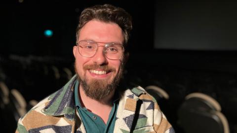 Hendrik Harms is interviewed in a cinema. There are seats behind him in the theatre with a screen in the background. He is wearing a green top and a jacket and wearing glasses. He has dark brown hair and a beard.