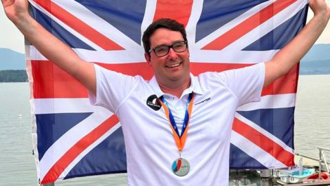 Karl Haines is holding the Union flag behind him whilst wearing his GBR Blind Sailing white polo shirt with a Silver Medal around his neck.