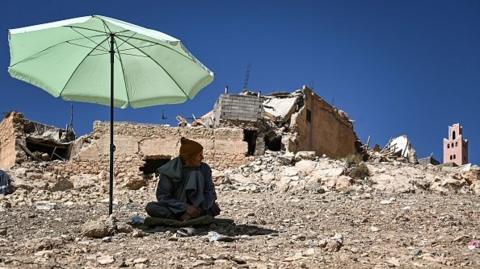 Man sits in shade of umbrella surrounded by rubble in Moulay Brahim, Morocco