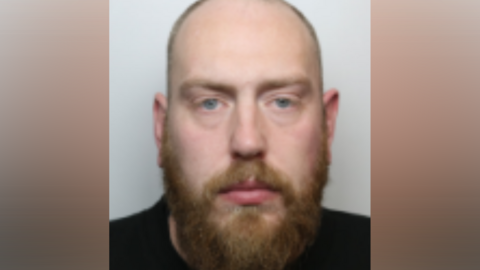 A police custody photo of Joseph Hewitt. He has receding red hair which is cropped short and a red beard. He is wearing a black top.
