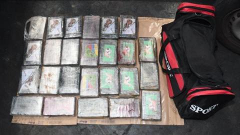 Multiple packages of suspected drugs, wrapped in cling film, are spread out on the floor on top of a flatten piece of cardboard. Beside the seized drugs is a large black and red gym bag.