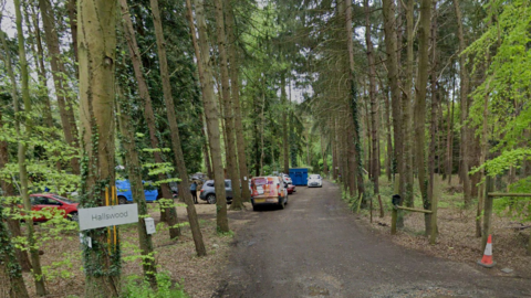 Hallswood sign beside a muddy track trailing out of sight, between trees, with a parked van and several cars visible. There is also a cone next to a tree.