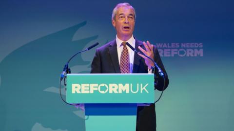 Nigel Farage, a man wearing a navy suit, stands at a podium with "Reform UK" on it, and a blue wall behind him.