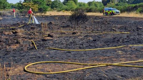 Two firefighters in red shirts and yellow trousers are holding long yellow hoses and spraying water onto scorched ground after a grass fire. In the background is a fire service vehicle parked on unburnt grass with trees and a blue sky behind.