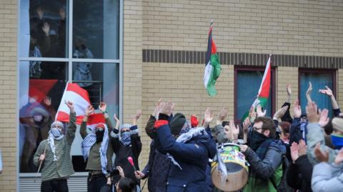 Protesters can been seen standing inside at the windows. Others are outside holding banners and flags.