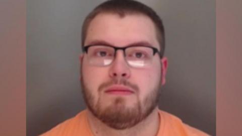 A mugshot of Sains looking into the camera. He has short, brown hair and a short beard. He is wearing rectangular glasses and an orange t-shirt.