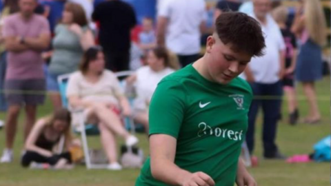 A young teen on a football pitch kicking a ball. His head is down and he is wearing a green football kit