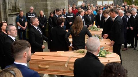 Three coffins in a row outside a church. There are flowers on each of them and people all around them, dressed in black.