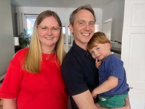 A family of three smiling, a mum with blonde hair, dad with grey and a little boy with ginger hiar cuddling his dad