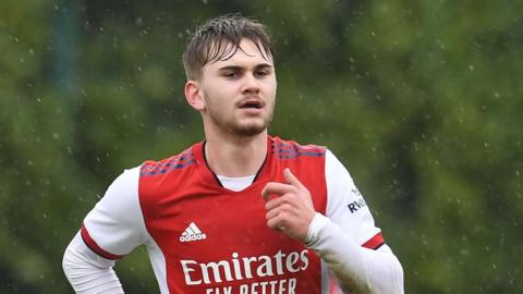 A young man with blonde hair wearing a red and white Arsenal football shirt while running in the run
