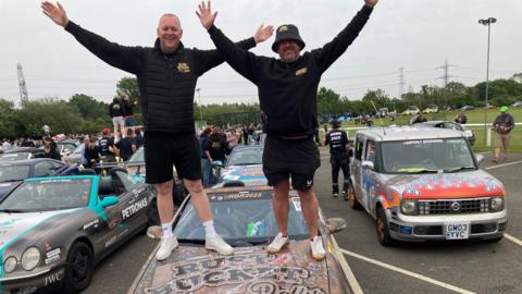 Two men, including Carlos Baldry, dressed in black long sleeved tops and shorts stand on the bonnet of a rust bucket rally vehicle with their arms aloft.