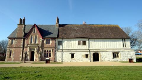 The image shows the Llanthony Secunda Priory building in bright sunshine. Two thirds of the building on the right is white walled and older looking, while the third on the left is made of red brick and more uniform. 