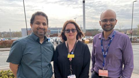 A woman and two men stand together in a hospital car park and smile at the camera. 