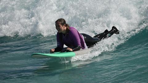 Kay Millar surfing on a wave. She is wearing a purple rash vest on top of a black wetsuit. She is surfing on a green and grey surfboard.