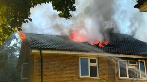 Flames can be seen coming through the roof of a badly damaged home. Water from firefighters is being sprayed towards the roof