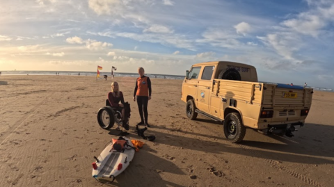 Wide angle photo of Perranporth beach. In the foreground of the photo on the beach there is a blonde woman who uses a wheelchair, she is facing the camera and smiling. Next to her is a person stood up wearing a wetsuit. On the sand in front of the people there is an adapted surfboard. A beige surf truck is parked next to the two people facing towards the sea, which can be seen in the distance.