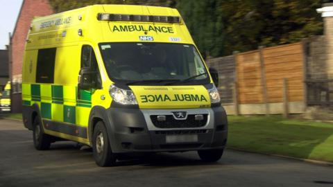 An ambulance vehicle drives along a road, fencing and a grass verge can be seen next to the vehicle.