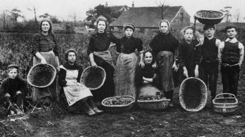 Black and white image showing ten girls and boys posing with baskets in front of crops on a farm, with farmhouses and trees in the background