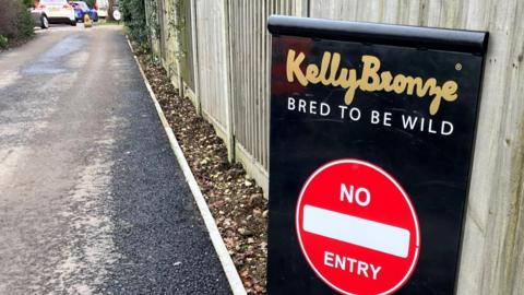 A black sign that says "KellyBronze, bred to be wild". Underneath the text is a red circle that says "no entry". The sign is at the bottom of a long driveway.