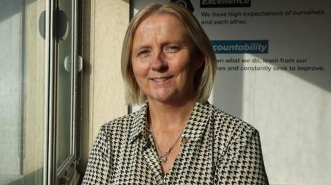 Rachael Hennigan standing in front of a window at the college. She has blond bobbed hair and is smiling and wearing a black and white dogtooth checked blouse