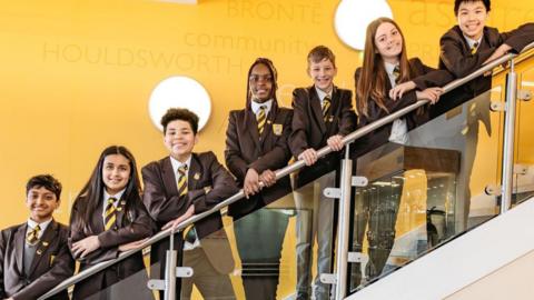 Seven pupils at Heckmondwike Grammar School, in brown blazers and trousers and yellow and brown striped ties, stand on stairs leaning on the handrail. The background wall is yellow. 