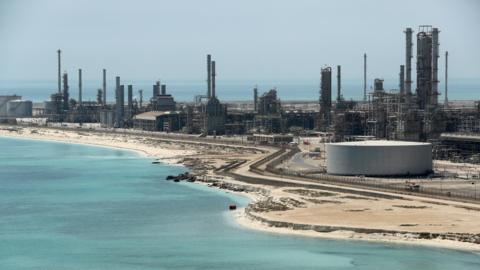 A file photo of Aramco's Ras Tanura oil refinery and oil terminal, which lies along a stretch of coast. The site comprises a network of several tall chimney-shaped structures and smaller buildings. It is daytime and the water along the coastline is a clear blue.