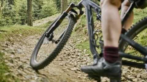 A man on a bike cycling along a rough track through woodlad. Only the man's legs are visible. 