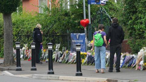 Four people stand with their backs turned outside Minehead Middle School. They are looking at bunch of flowers which have been lined up against the metal fence outside the school. There is a red balloon in the shape of a heart attached to the fence.