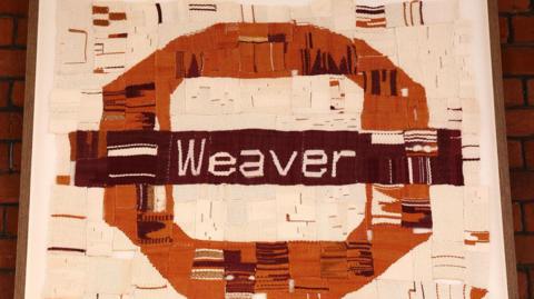 A woven London Overground Weaver line roundel in maroon, orange, and cream, displayed on a red brick wall at Hackney Downs station.